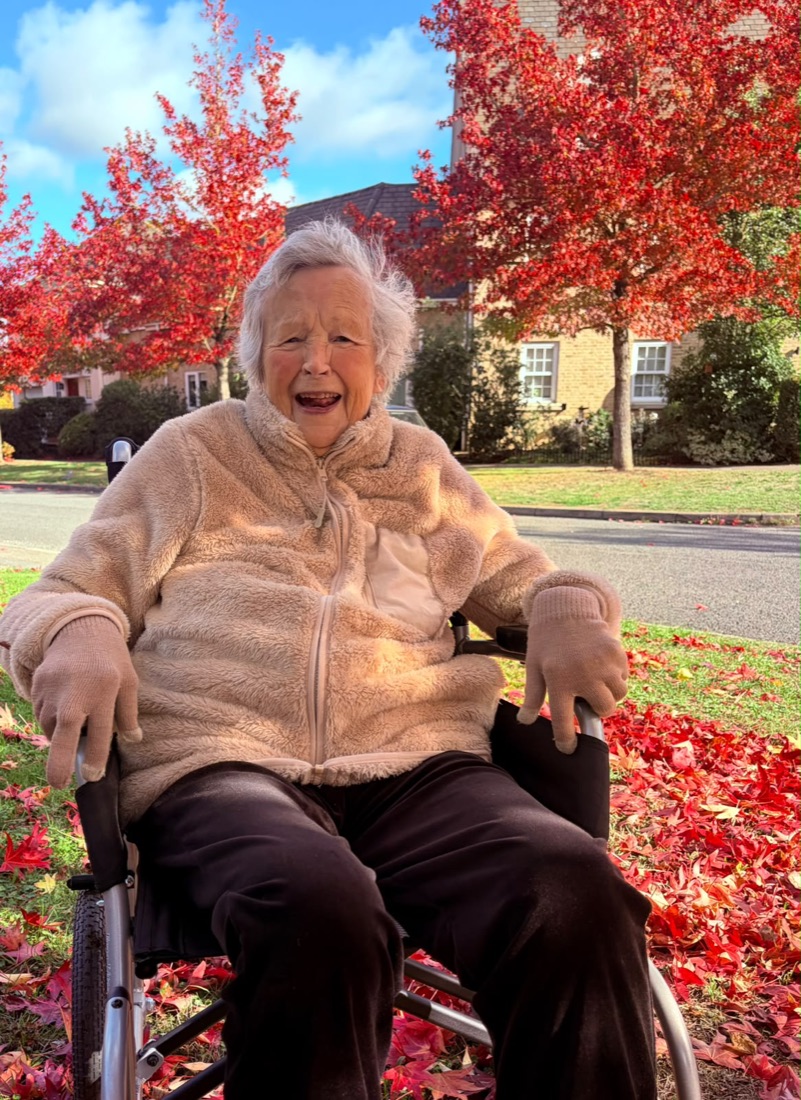 Josie laughing in her wheelchair under bright red autumn trees on a sunny day